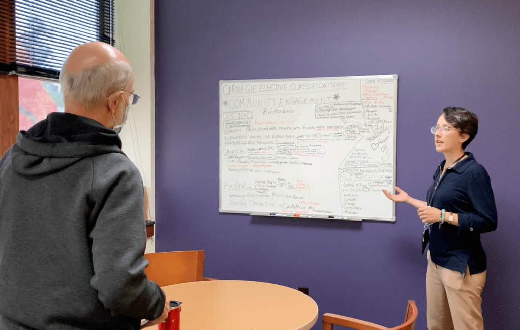 A young woman stands in front of a white board that is full of writing as she speaks to an older man